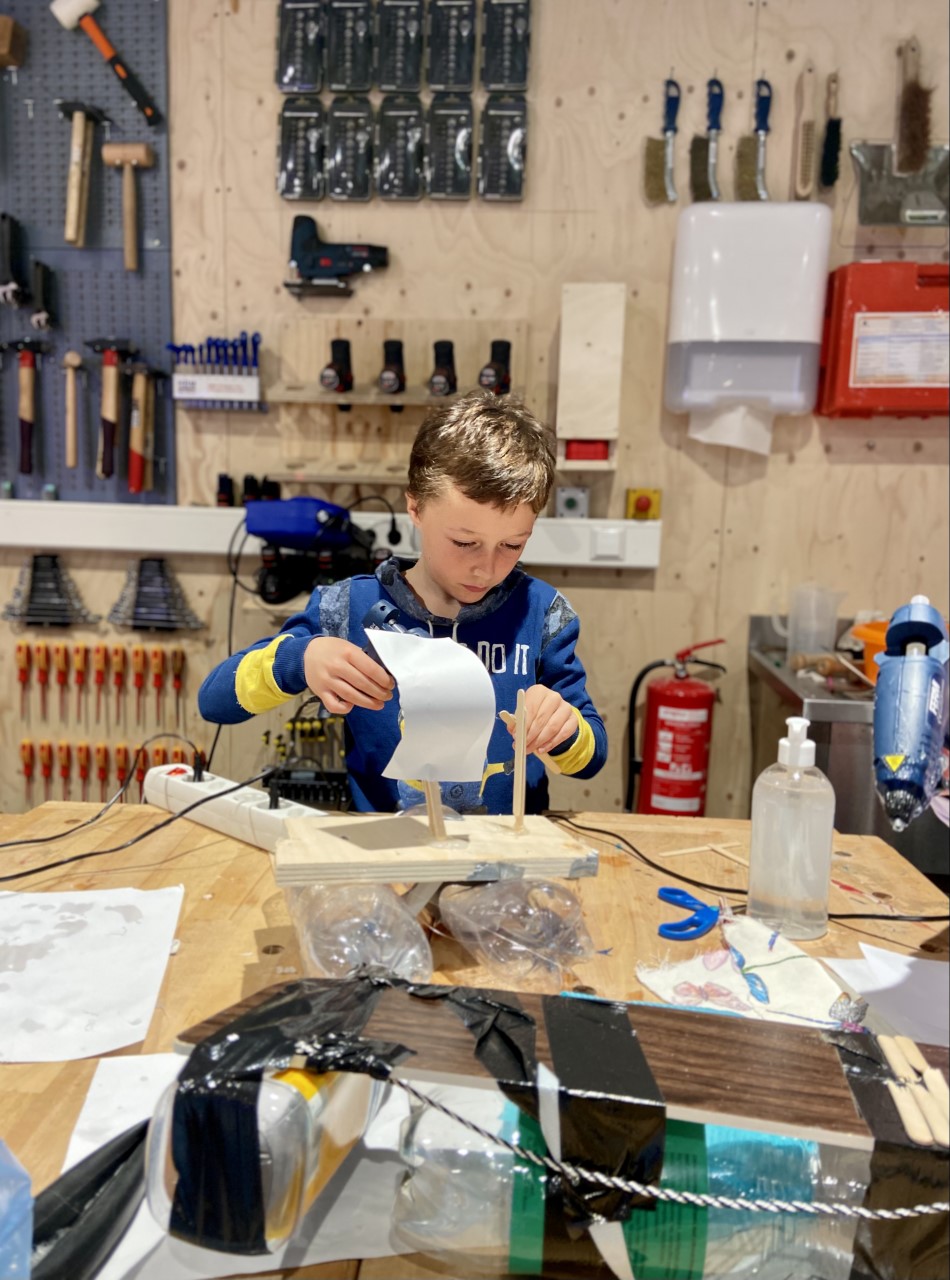 Een gefocuste jongen die aan het werken is met hout en papier, hij staat voor een grote gereedschapsmuur vol met tangen, boren, en hamers.